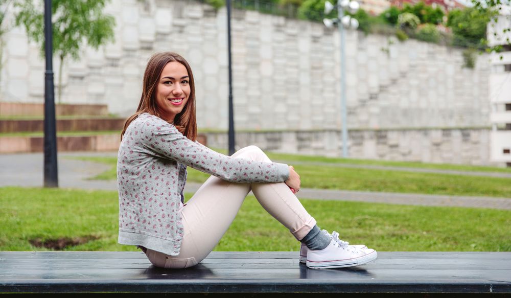 Side of young woman sitting outside on park bench smiling at camera 