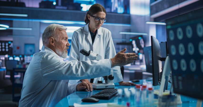 Male Neuroscientist Using Microscope to Analyze Petri Dish Sample And Talking To Female Docor In Medical Research Center.