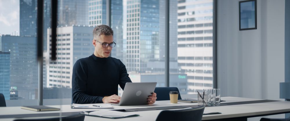Businessman Sitting Down At His Desk And Starts Typing On Laptop Computer In Corner Office With Megapolis Cityview.