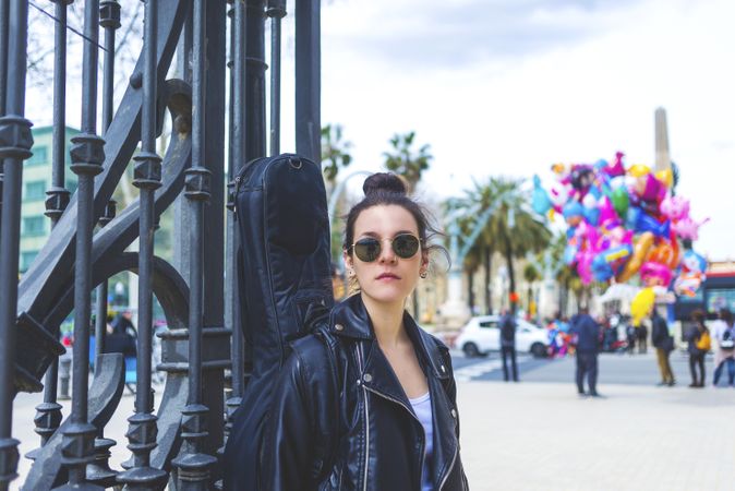 Front view of a young laughing hipster woman standing and leaning on a fence at park in sunny day while looking away