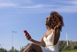 Excited female with curly hair sitting on park grass talking on phone
