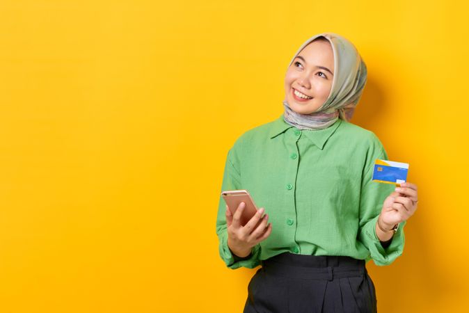 Smiling Muslim woman in headscarf with credit card and smart phone looking up