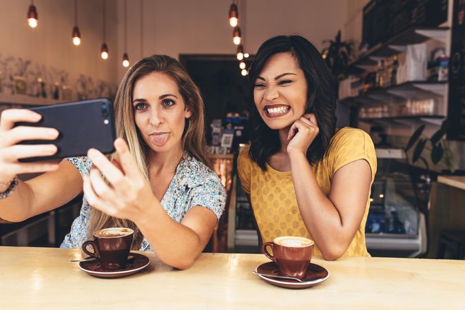 Woman sticking out her tongue while taking selfie with a smiling friend at coffee shop