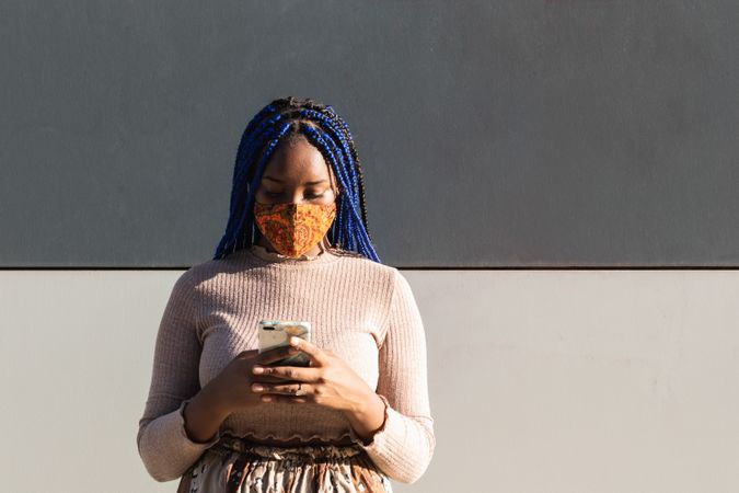 Black woman in mask messaging on smartphone in city