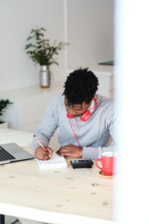 Black student with headphones writing in notebook at home