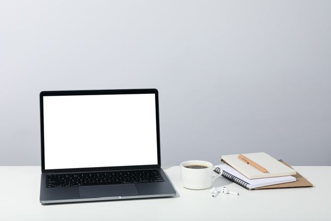 Table with open laptop with mockup screen, earbuds, coffee, and blank notebooks, with copy space