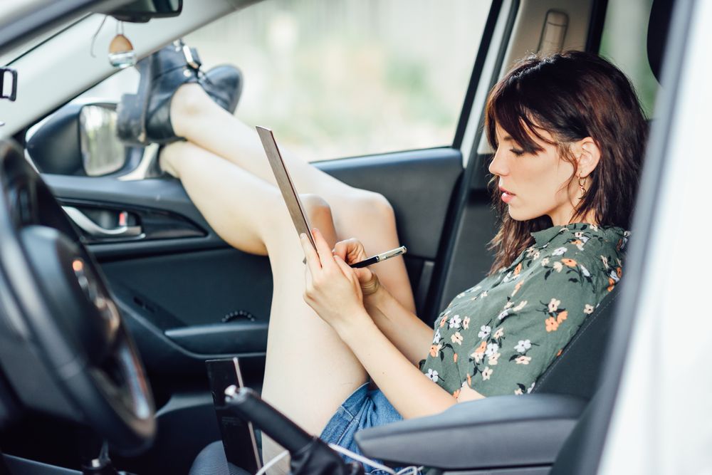 Female wearing floral shirt and shorts writing notes while sitting in parked car