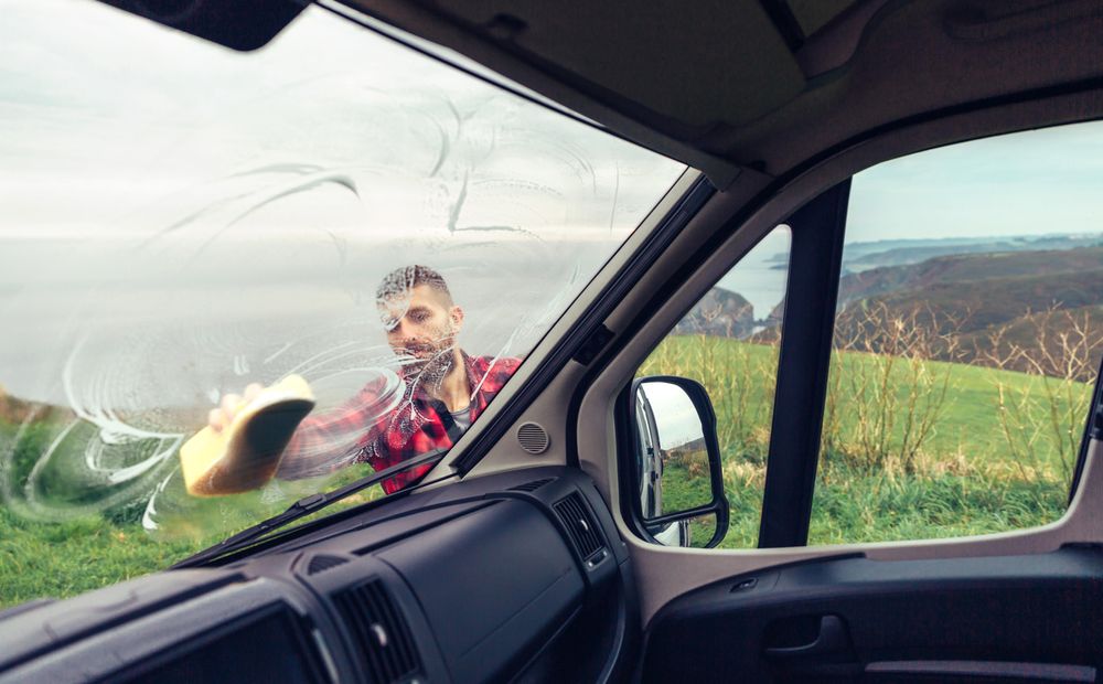 View from inside van of man in checkered red shirt cleaning front window with sponge