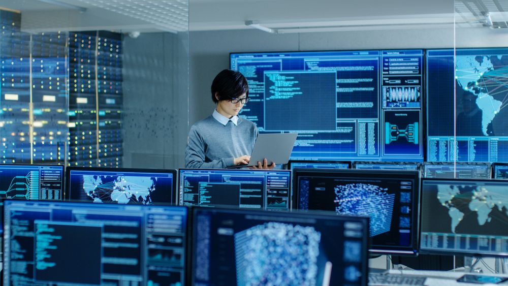 In the System Control Room IT Technician Holds and Works on a Laptop, in Background Multiple Displays with Graphics.