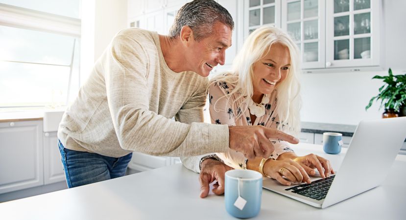 Older couple laughing and smiling while typing on a laptop in the kitchen