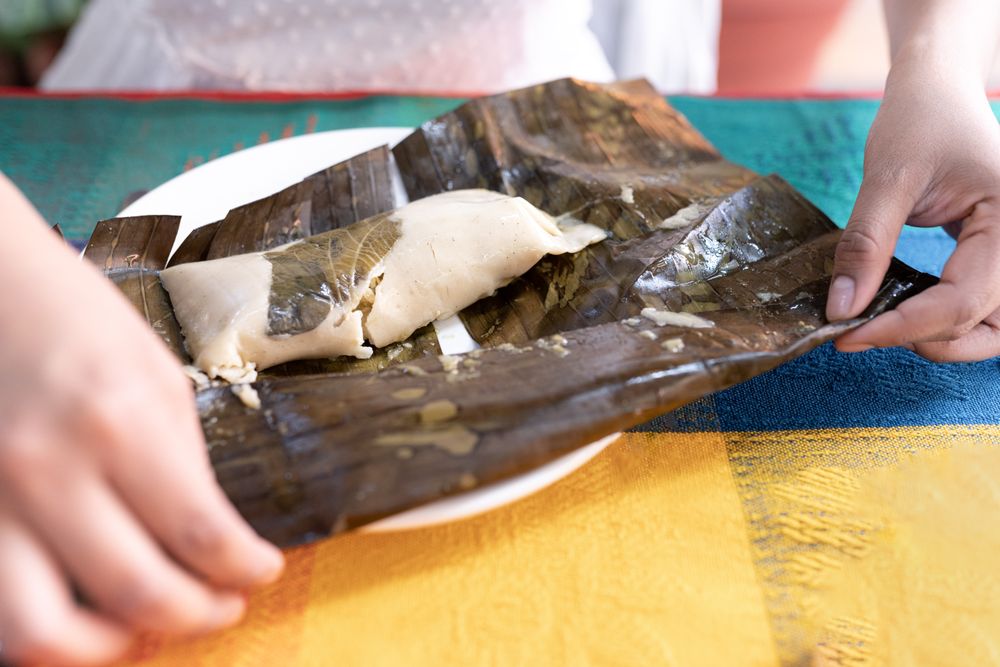 A young Hispanic woman is opening a meat tamale wrapped with a plant leaf in a restaurant