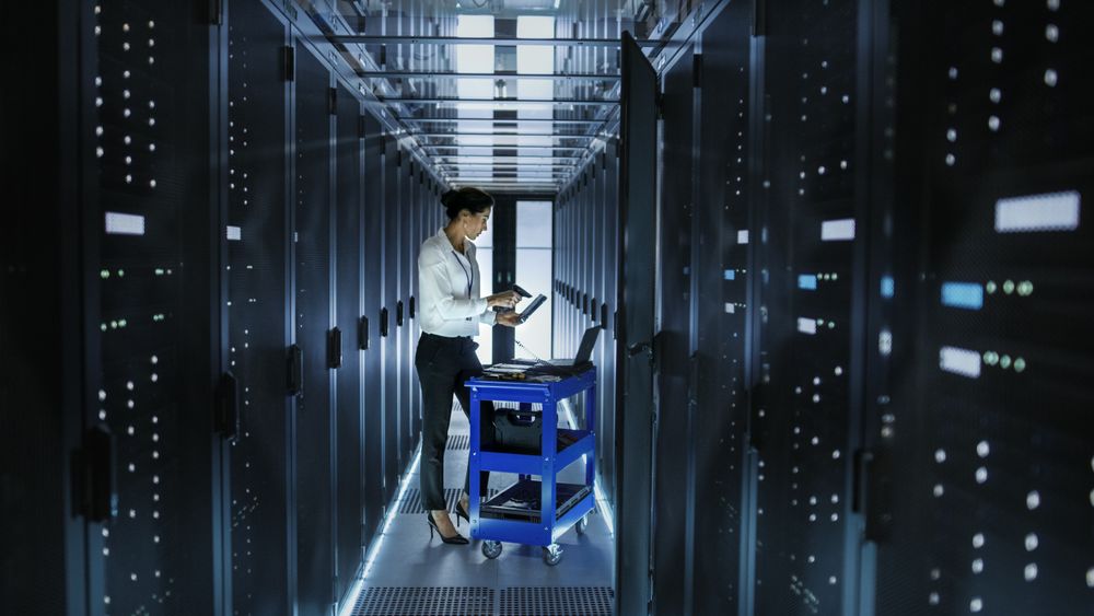 Female IT Engineer Works on a Laptop and Scans and Swaps Hard Drives That are Lying on a Cart.