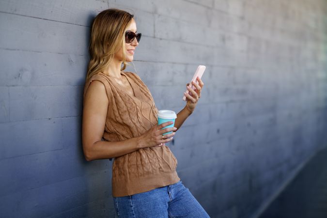 Woman with coffee leaning on wall and using smartphone