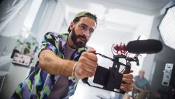 Young Male Videographer With Beard, Wearing Colorful Shirt And Headband, Operates Camera With Microphone In Studio Setting, Capturing Creative Content For Advertising Or Movie Production.