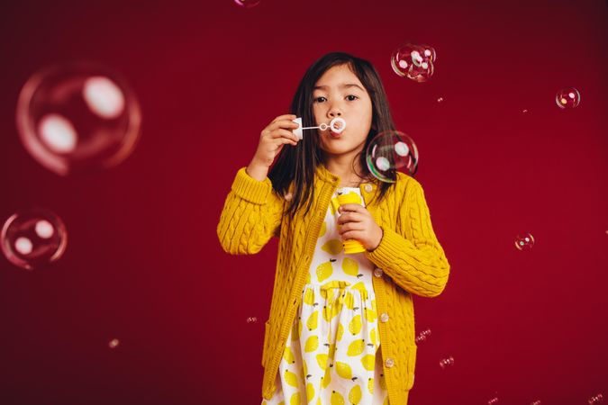 Little girl standing against a red background blowing bubbles
