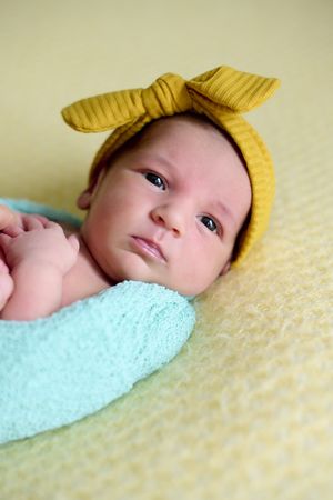 Close-up of a newborn baby girl, inside a ball of blue wool on a yellow blanket with a yellow bow on her head