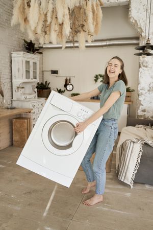 Joyful strong woman carrying washing machine in kitchen