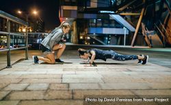 Woman doing push ups with support from personal trainer in the city at night 4AzmE6
