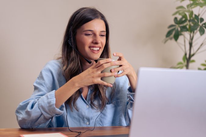 Young female professional enjoying coffee during virtual meeting wearing headset and working remotely with laptop in home office