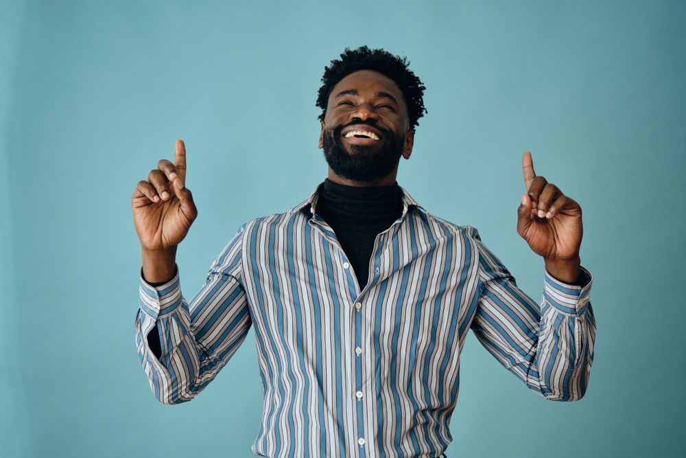 Smiling Black man in striped shirt and jeans pointing up with both hands with his head back