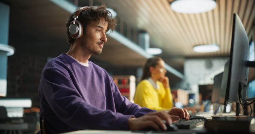 Portrait of a Stylish Male Student Working on His University Project on a Desktop Computer. Young Handsome Man Studying a Course Online in a Quiet Public Library with Bookshelves with Books