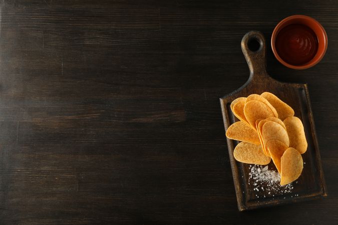 Potato chips on a wooden board with salt on a dark background