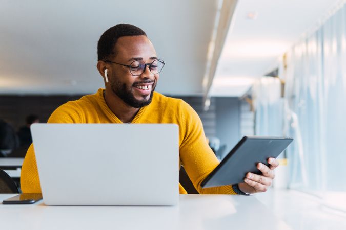 Cheerful man with earbuds using tablet and laptop at table