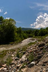 Rocky riverbank with flowing stream, surrounded by lush greenery and distant hills.