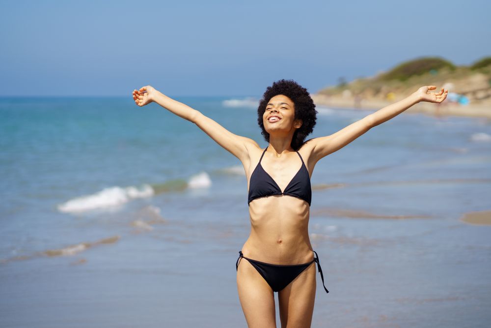 Smiling woman in bikini standing on beach with outstretched arms and eyes closed