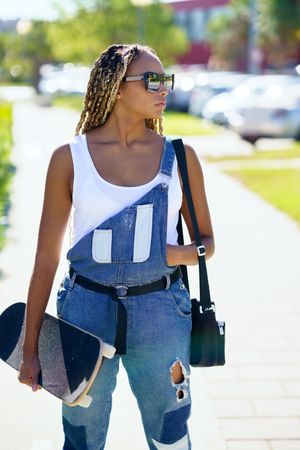 Female in denim overalls standing with skateboard and looking away