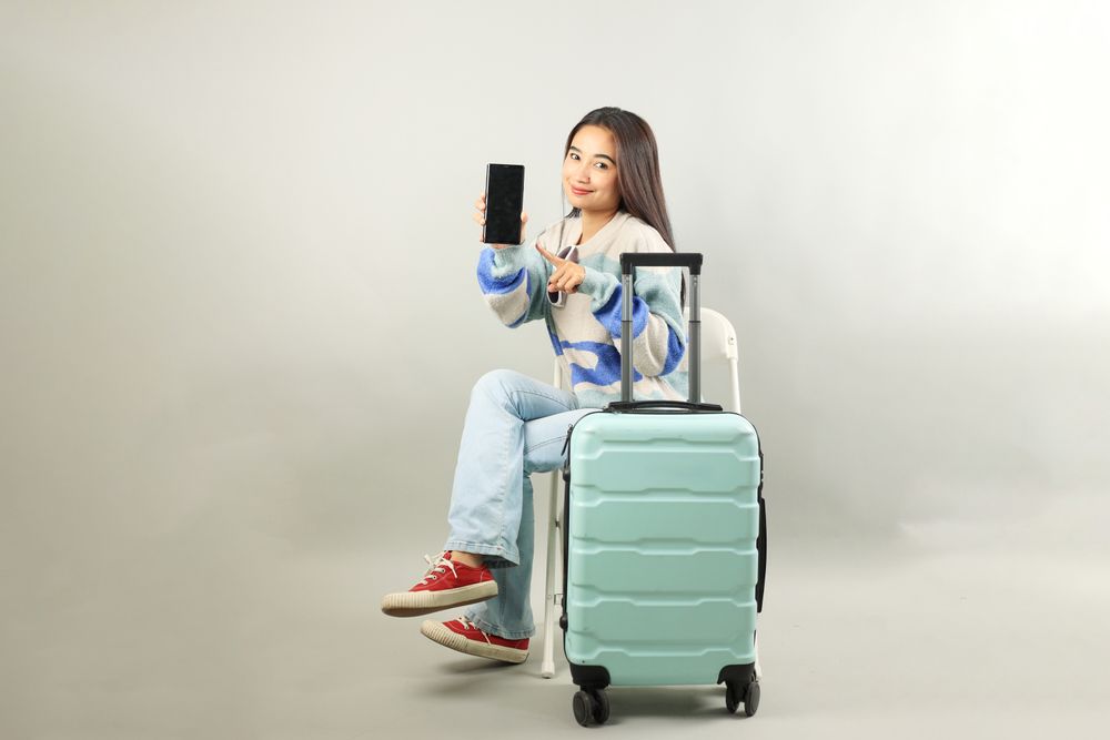 Happy Young Asian Woman Sitting on Chair with Suitcase and Showing Mobile Phone Screen