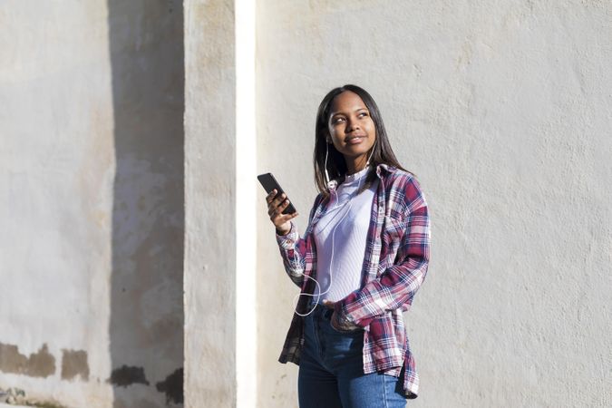 Female standing in the sun in front of wall holding phone