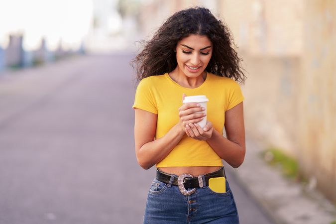 Shy female in yellow t-shirt walking on street with coffee, copy space