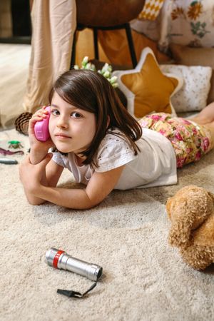 Little girl talking with toy phone lying over rug surrounded by cushions and plants in teepee