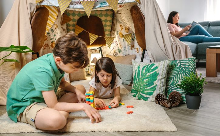 Children playing together with stacking piece game while relaxed mother reading book sitting on sofa