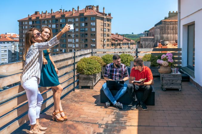Two female friends taking a selfie in rooftop while their colleagues working with laptop