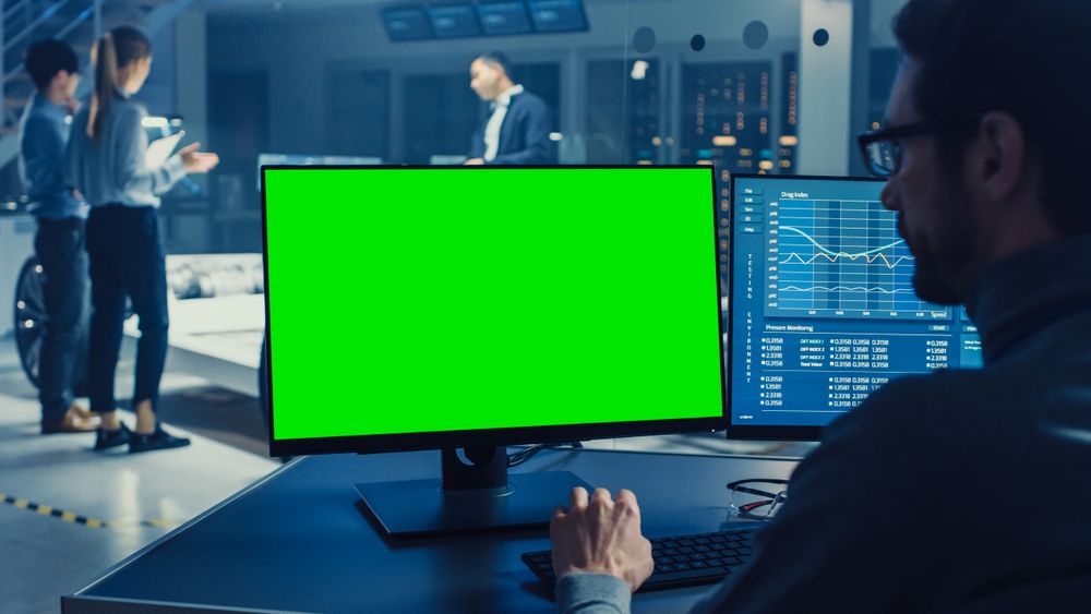 Professional Engineer Works on a Computer with Green Screen Mock Up and Tests the Electric Car Chassis Prototype with Wheels, Batteries and Engine Standing in a High Tech Development Laboratory.