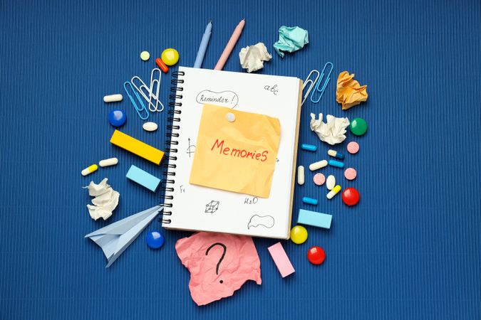 “Memories” post it note on book with pills, paper clips and pens on blue background