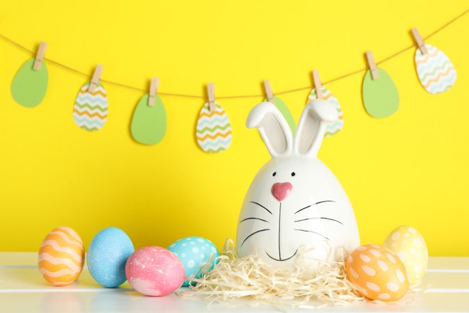 Decorated bunny and Easter eggs on table against color background