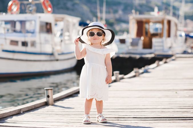 Smiling girl in light dress with hat and sunglasses standing beside docked boats