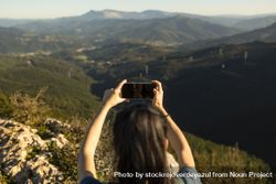 A happy young woman takes pictures of the landscape with her smartphone from the top of a mountain bYqq36