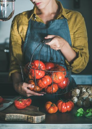 Woman in yellow holding wire basket of large tomatoes in kitchen