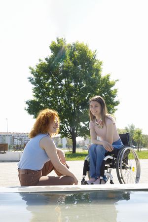 Woman in wheelchair and red haired friend sitting in front of a park fountain