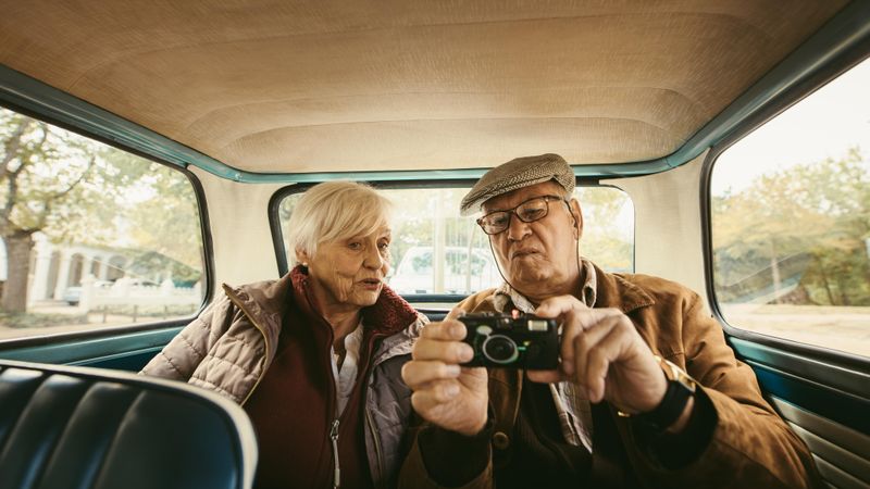 Older married couple sitting on backseat of car and looking at digital camera