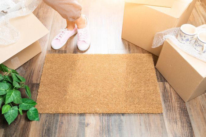 Woman in Pink Shoes and Sweats Standing Near Home Sweet Home Welcome Mat, Boxes and Plant