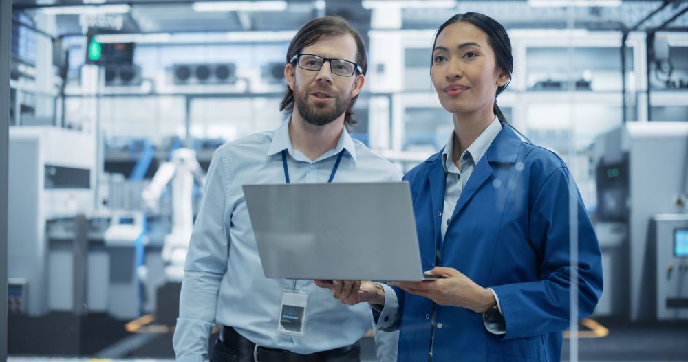 Industrial Engineers Working at a Technology Factory. Man and Asian Female Using Laptop Computer and Having a Conversation. Footage with a Portrait of a Couple of Employees at a Plant