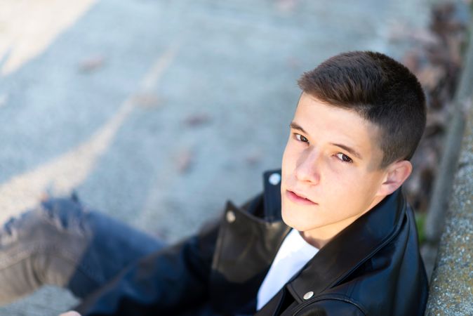 Teenage male wearing a leather jacket leaning on ledge outside looking up at camera