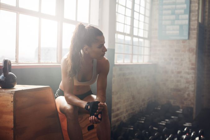 Indoor shot of healthy young woman relaxing on box at gym and looking away