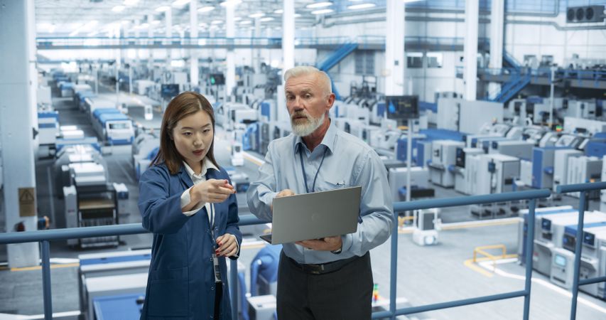 Mature Engineer and Young Japanese Technician Having a Conversation and Using Laptop Computer. Specialists Monitoring, Analyzing Conditions at a Modern Electronics Factory with Automated Robots