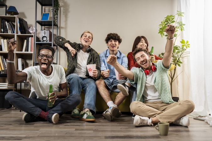 Group of diverse young friends cheering and eating popcorn together during a fun living room movie night